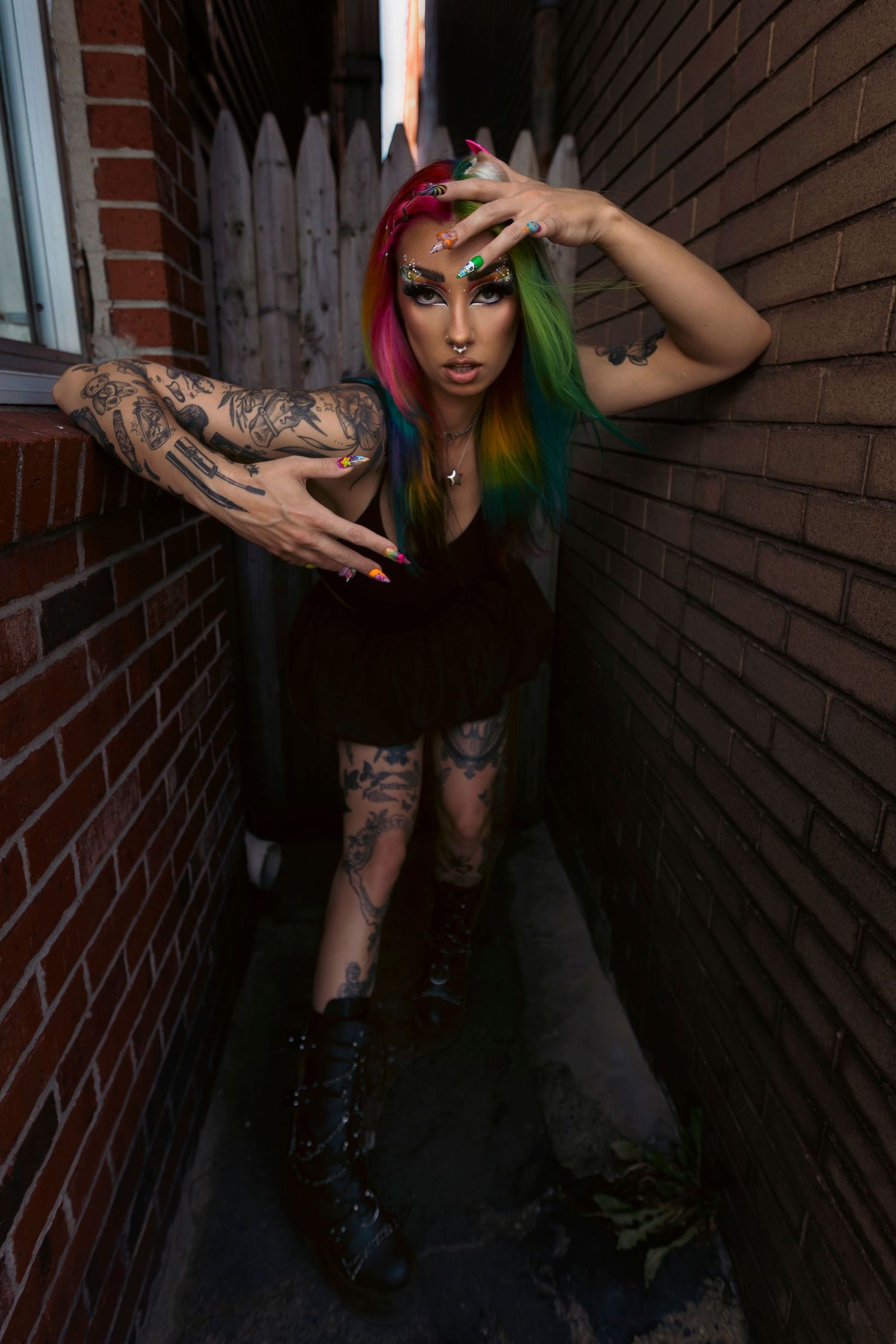 Alternative model with vibrant red and green hair poses dramatically in a narrow brick alley wearing dark attire and boots.