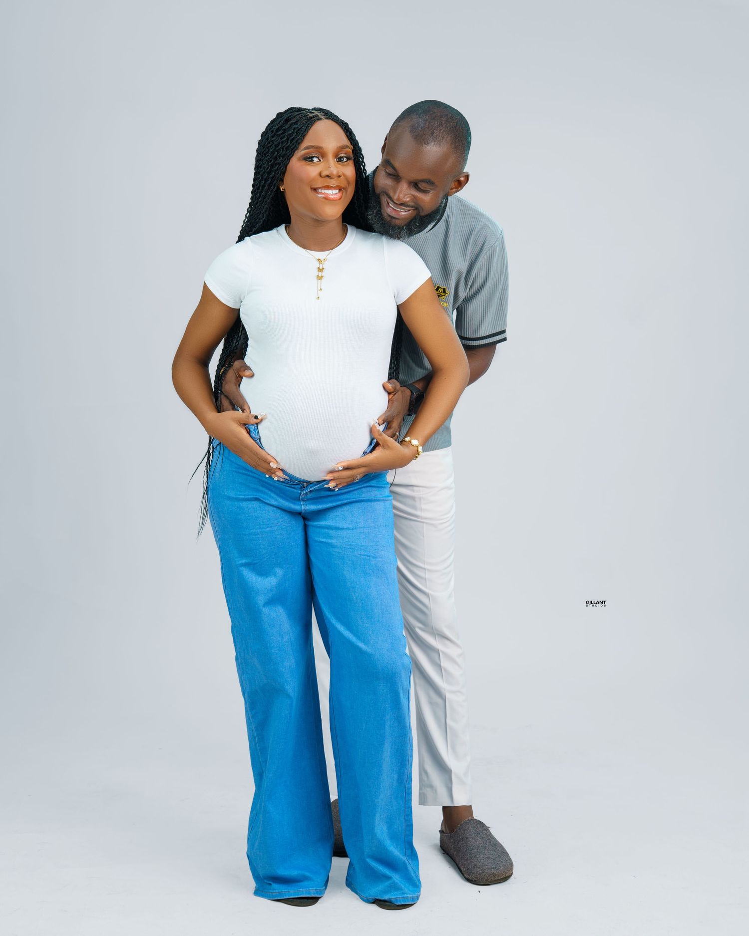 Expectant couple embracing in casual wear against light gray background during maternity photoshoot.