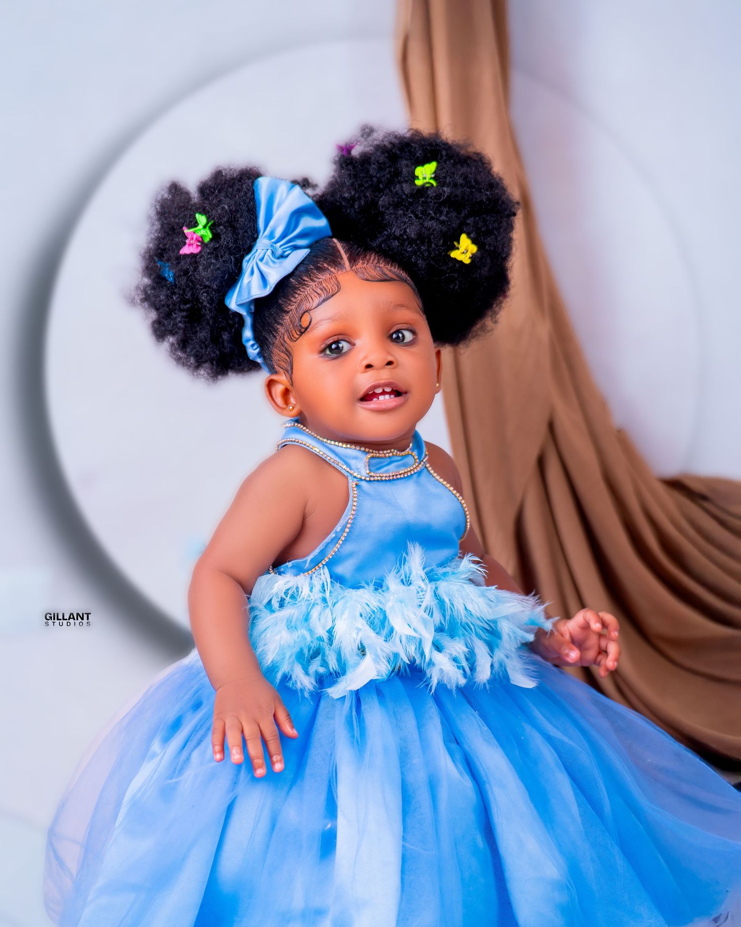 Small child in a flowing blue tulle princess dress with butterfly accessories in their natural hair against brown backdrop.