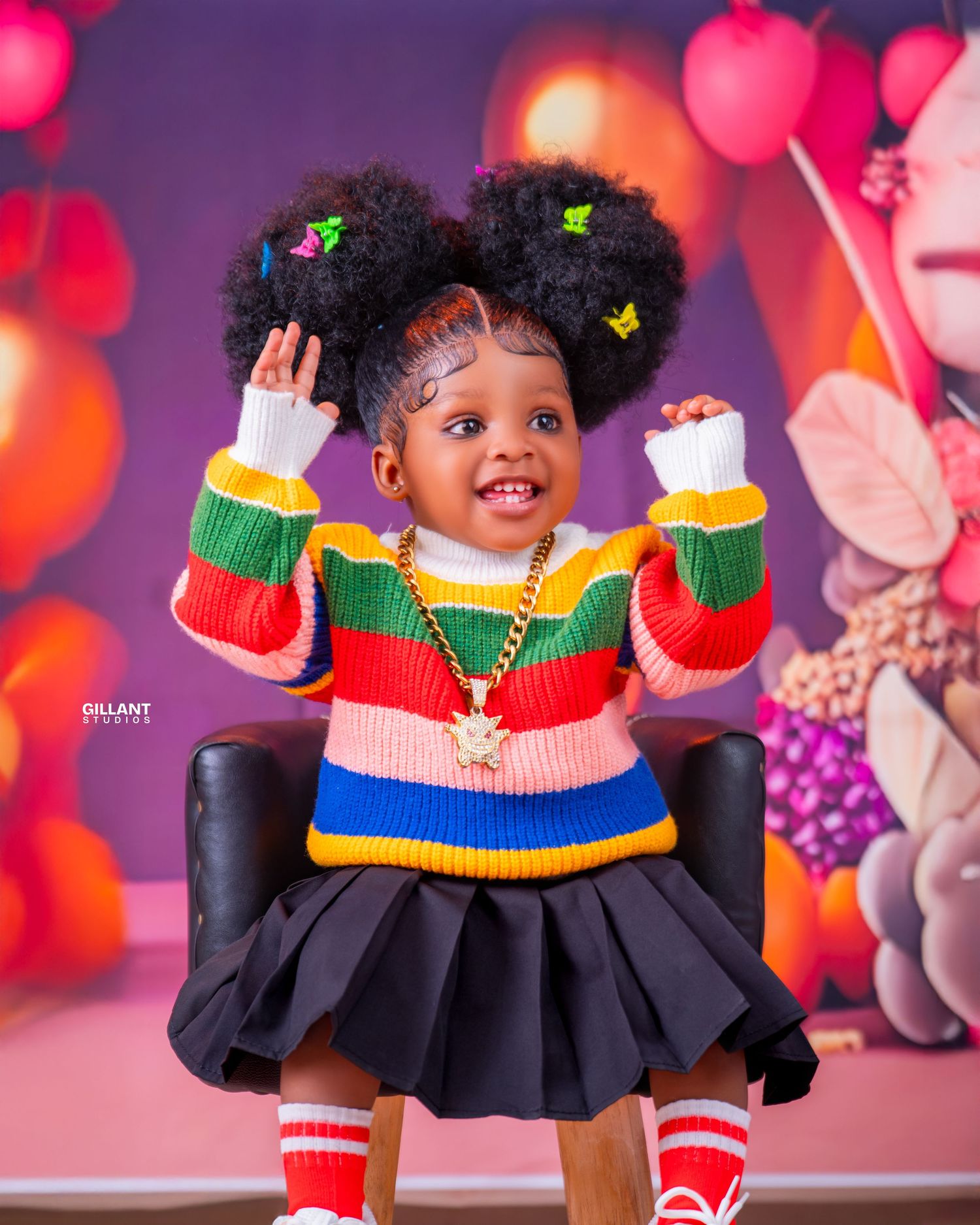 Joyful toddler in striped sweater and skirt poses with hands up amid festive balloon backdrop.