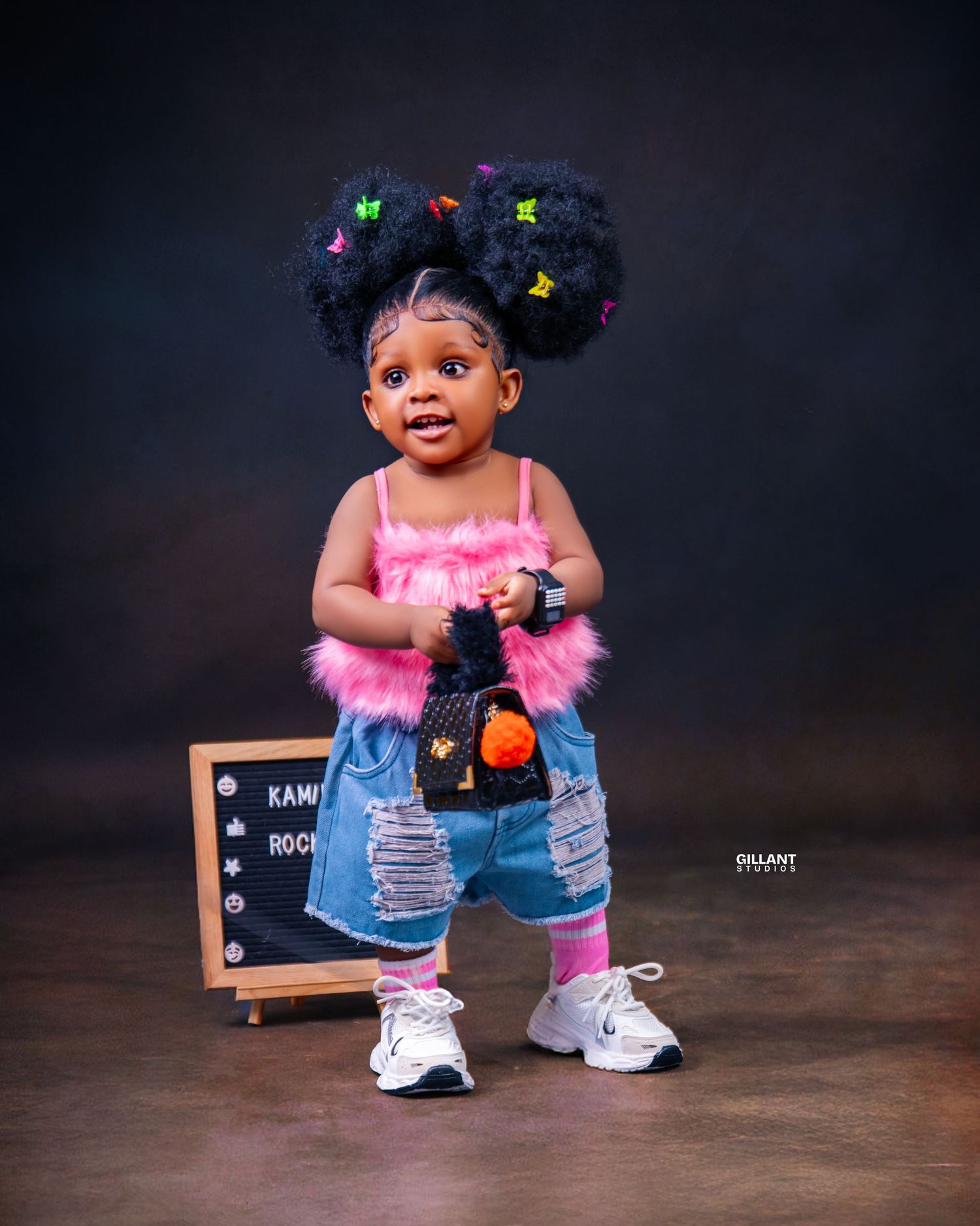 Toddler in pink top and distressed denim with natural hair adorned with butterflies poses by chalkboard.