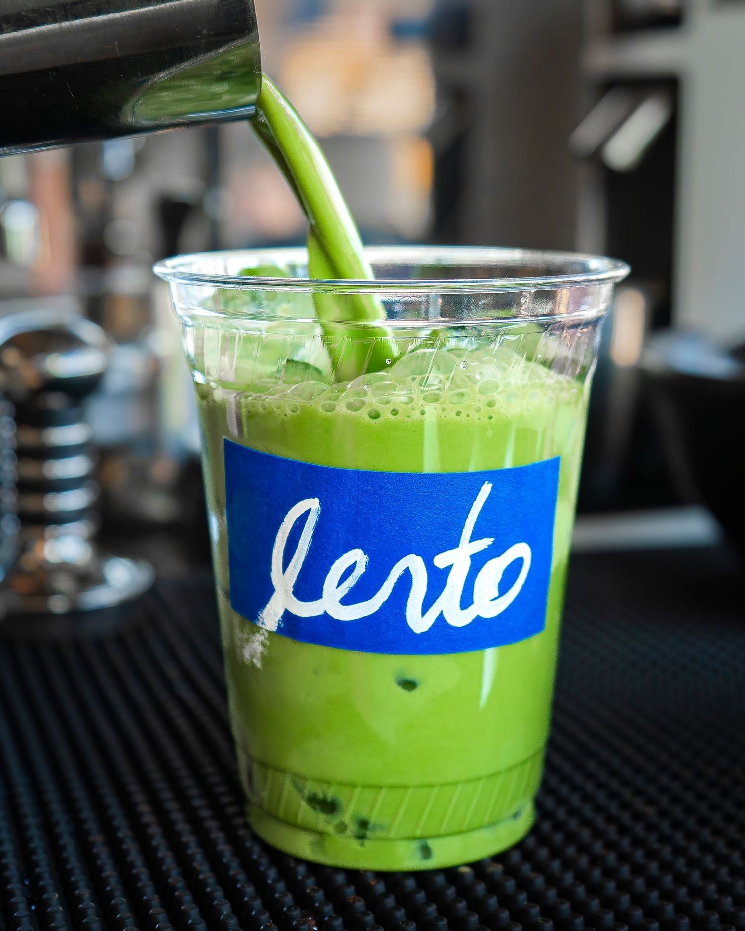 Iced matcha drink being poured into a clear cup labeled “Lerto” on a bar counter.