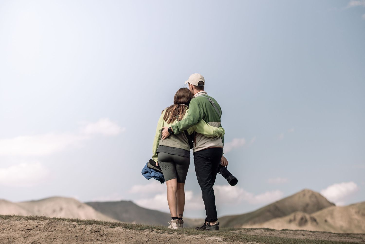 Two people hiking together along a scenic mountain trail with rolling hills and distant peaks in view.