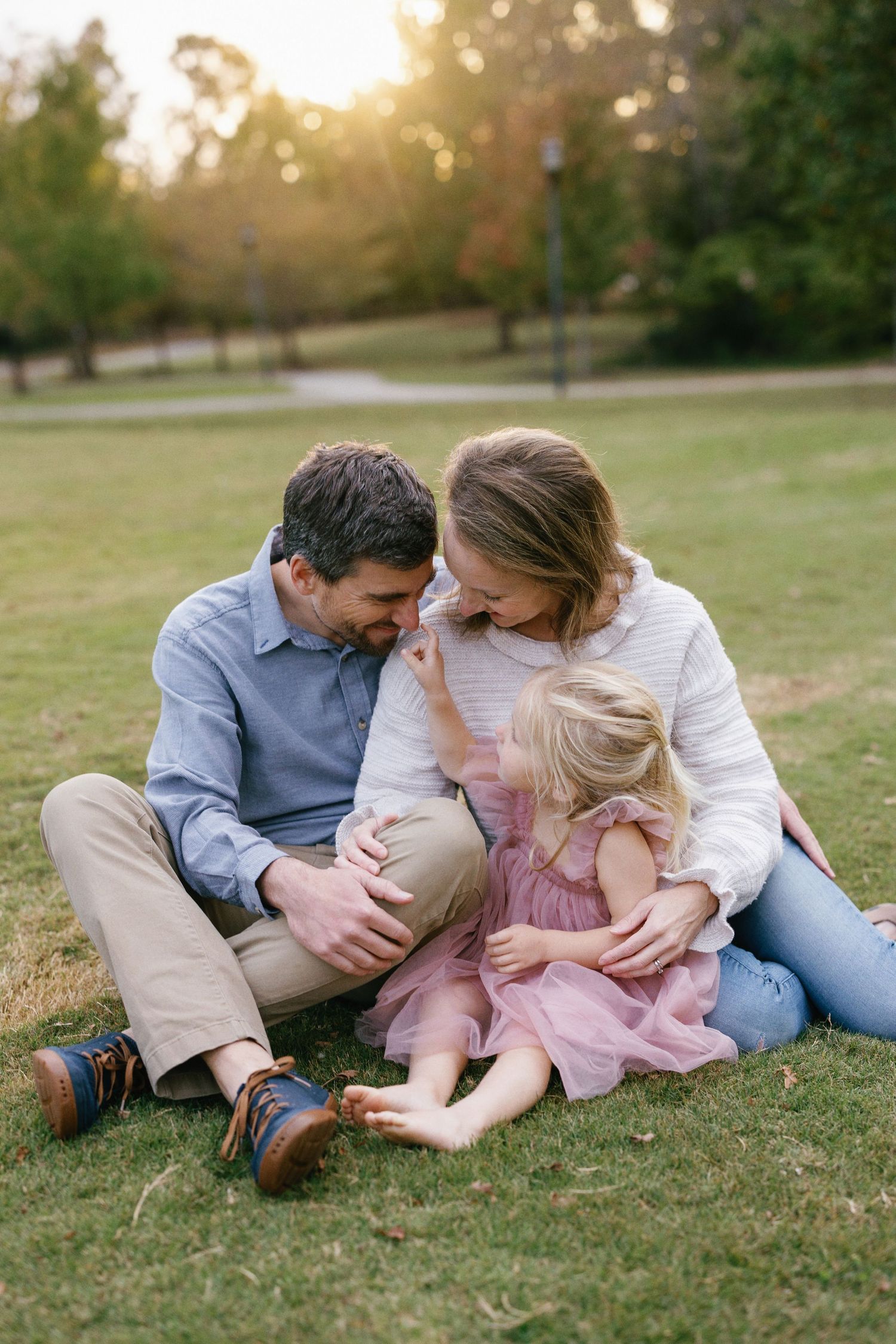 A family lies together on grass during golden hour while sharing tender playful moments in a park.