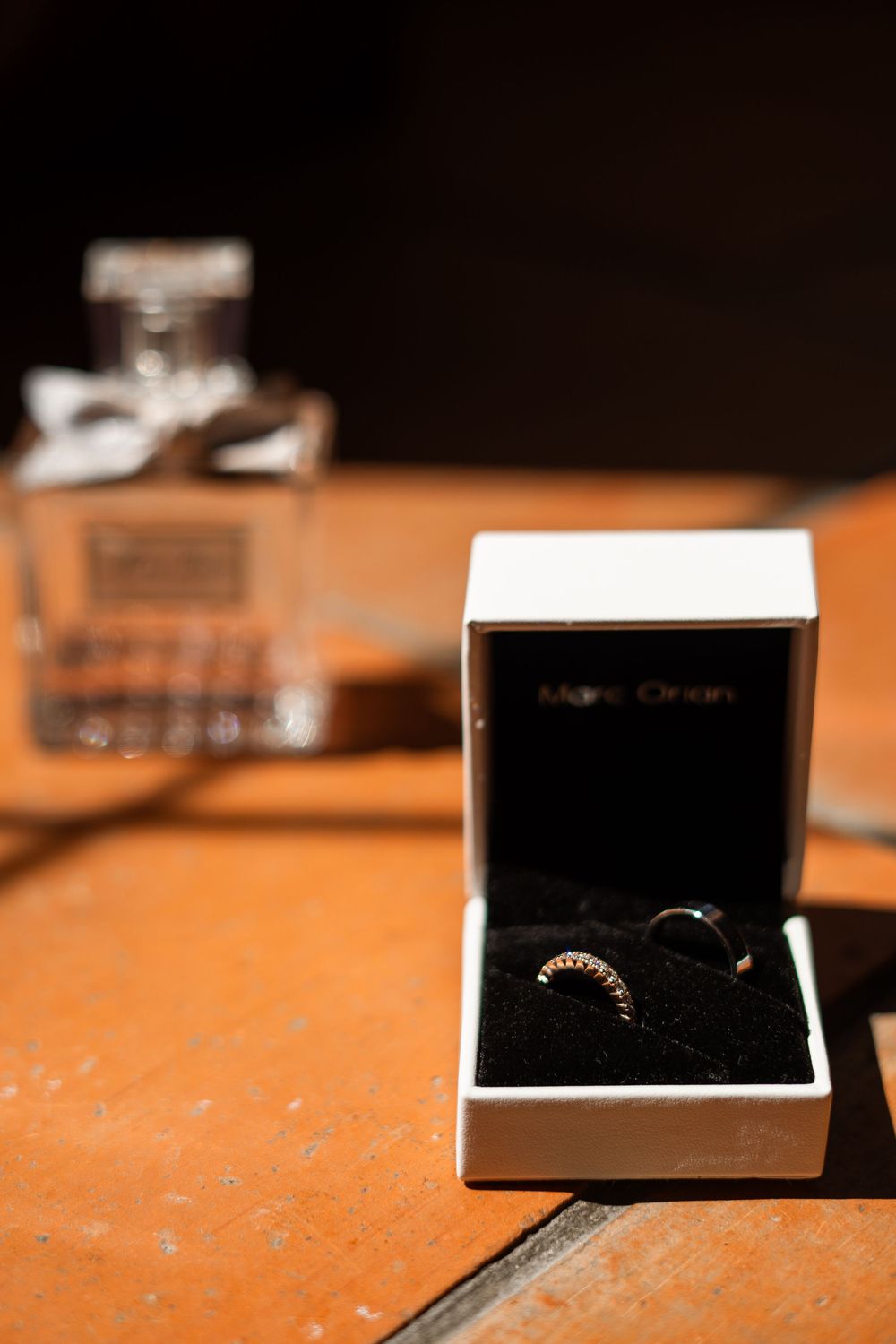 A small black jewelry box with rings inside sits on a wooden surface with a perfume bottle in the background.
