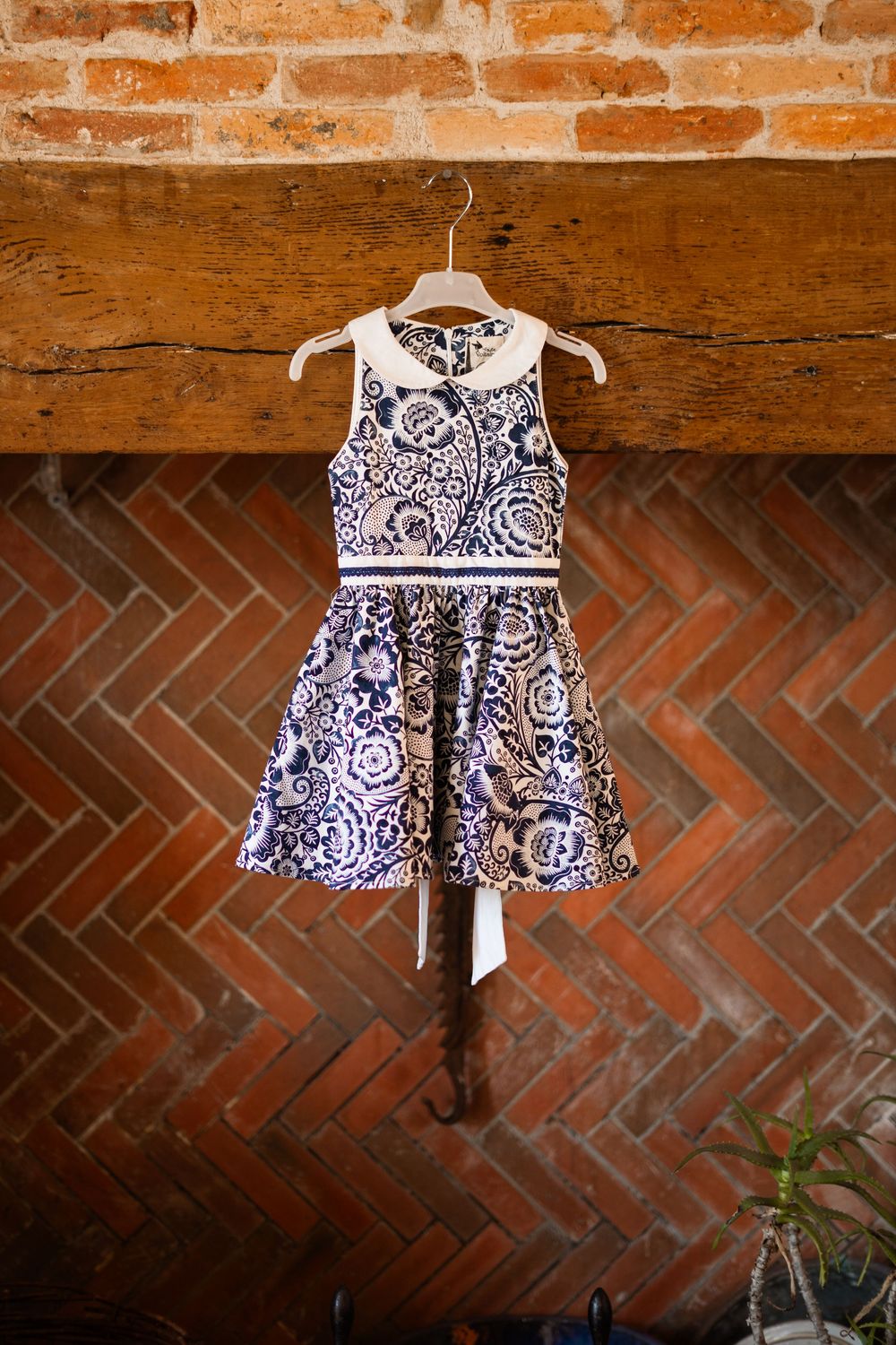 A blue and white floral print flower girl dress hangs against an exposed brick wall.