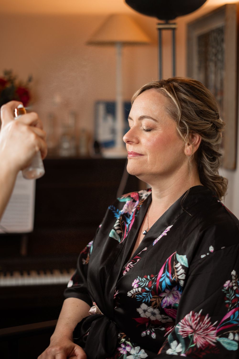 Having makeup applied while wearing a floral robe and relaxing with eyes closed beside a piano in warm lighting.