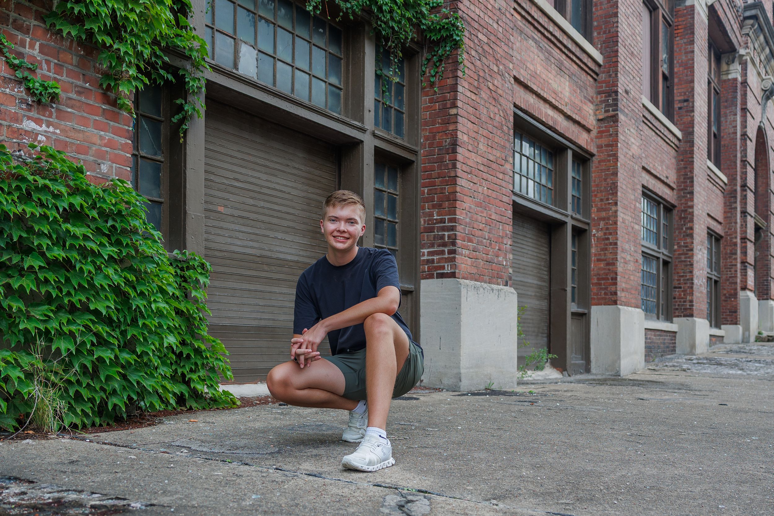 Urban brick building exterior with ivy-covered walls and concrete steps. Senior boy posing in a crouched down position.