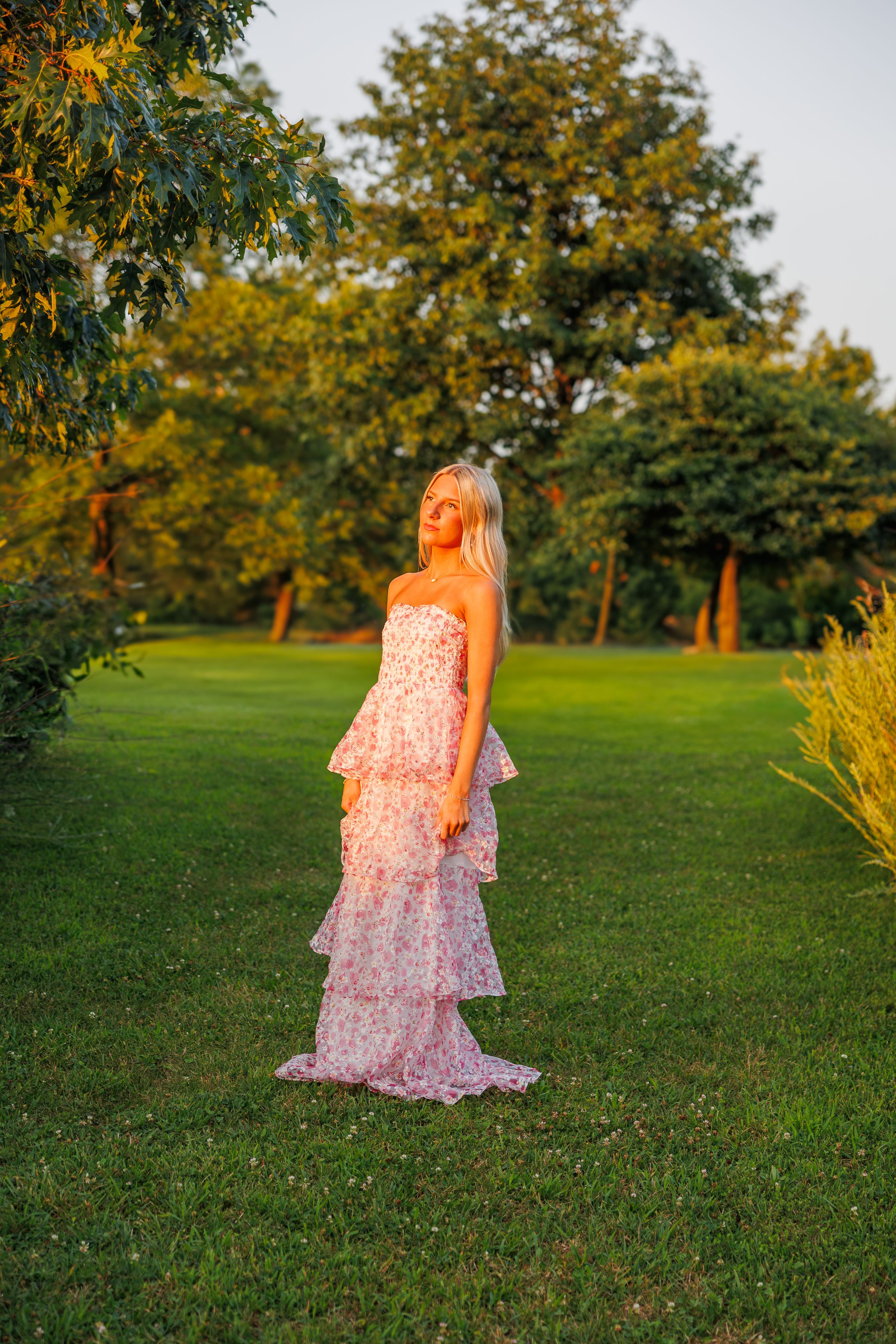Flowing pink dress photographed in a sunlit grassy field during golden hour. Senior girl posed in golden hour sunlight.