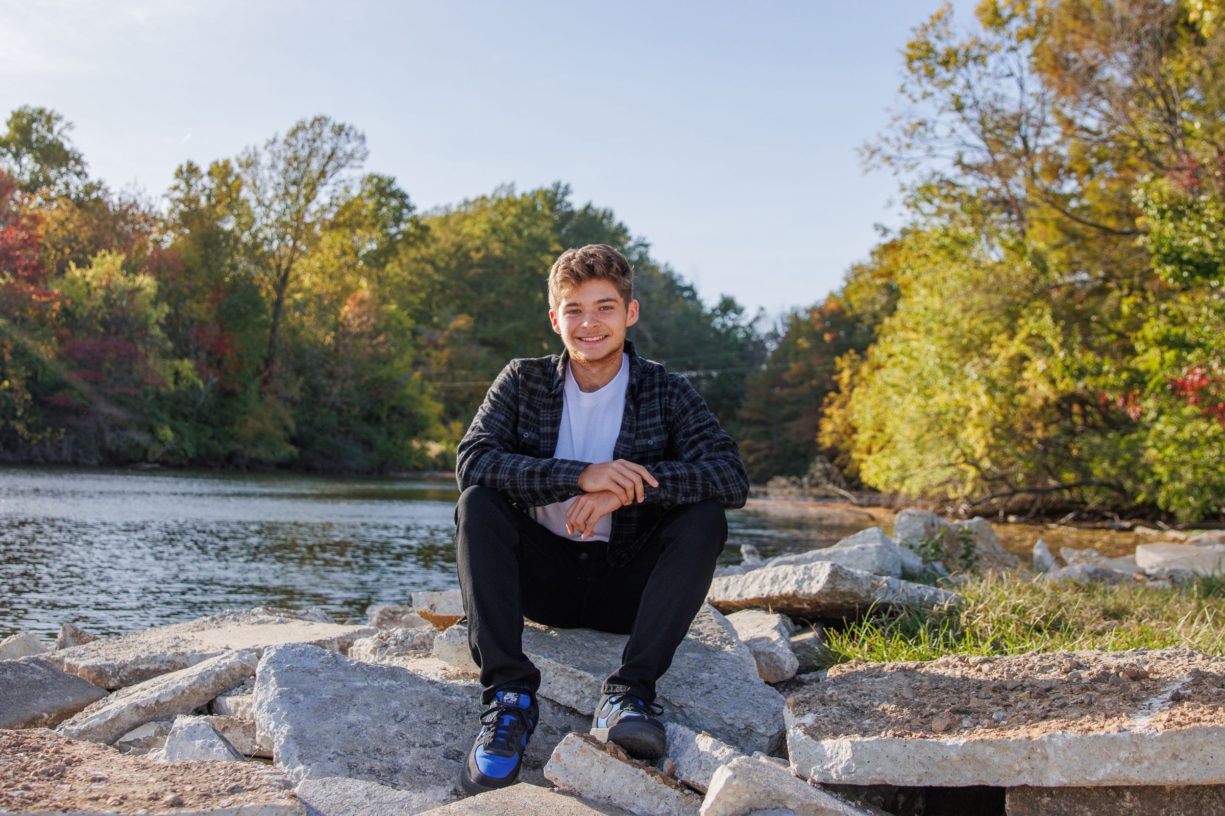 Scenic autumn riverside photoshoot showing rocky terrain and fall foliage. Senior boy sitting on rocks near the lake.