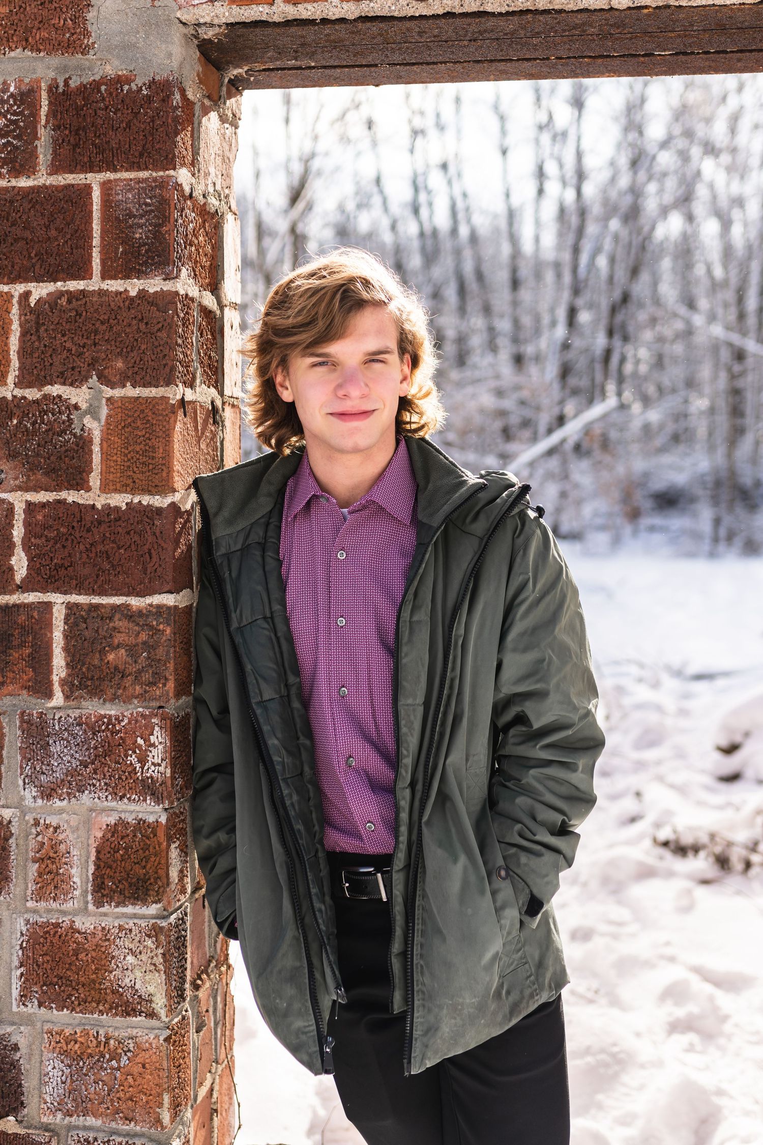 Winter photo of a senior boy leaning against a brick wall with snow covered trees behind him.