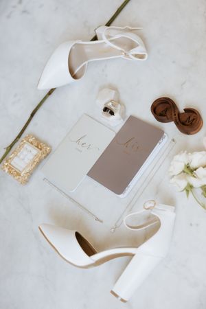 Elegant wedding flat lay with white heels, his and hers vow books, ring box, perfume, and floral accents on marble.