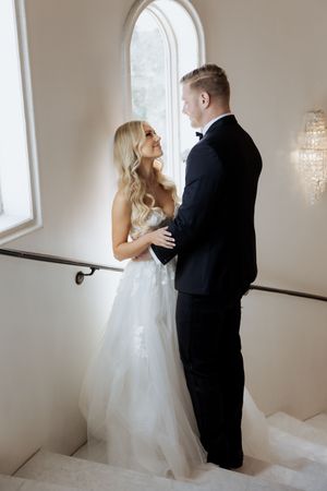 Bride in a white tulle gown and groom in a black tuxedo share a romantic moment on a grand staircase.