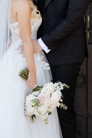 Bride in lace wedding gown holding white and blush bouquet stands beside groom in black suit near ornate dark doors.