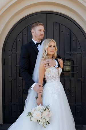 Bride and groom pose elegantly before ornate black arched doors, bride holding white floral bouquet in lace ball gown.