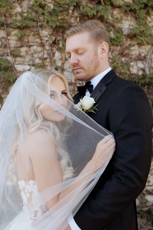 Romantic bride and groom portrait with flowing veil against ivy wall in wedding attire.