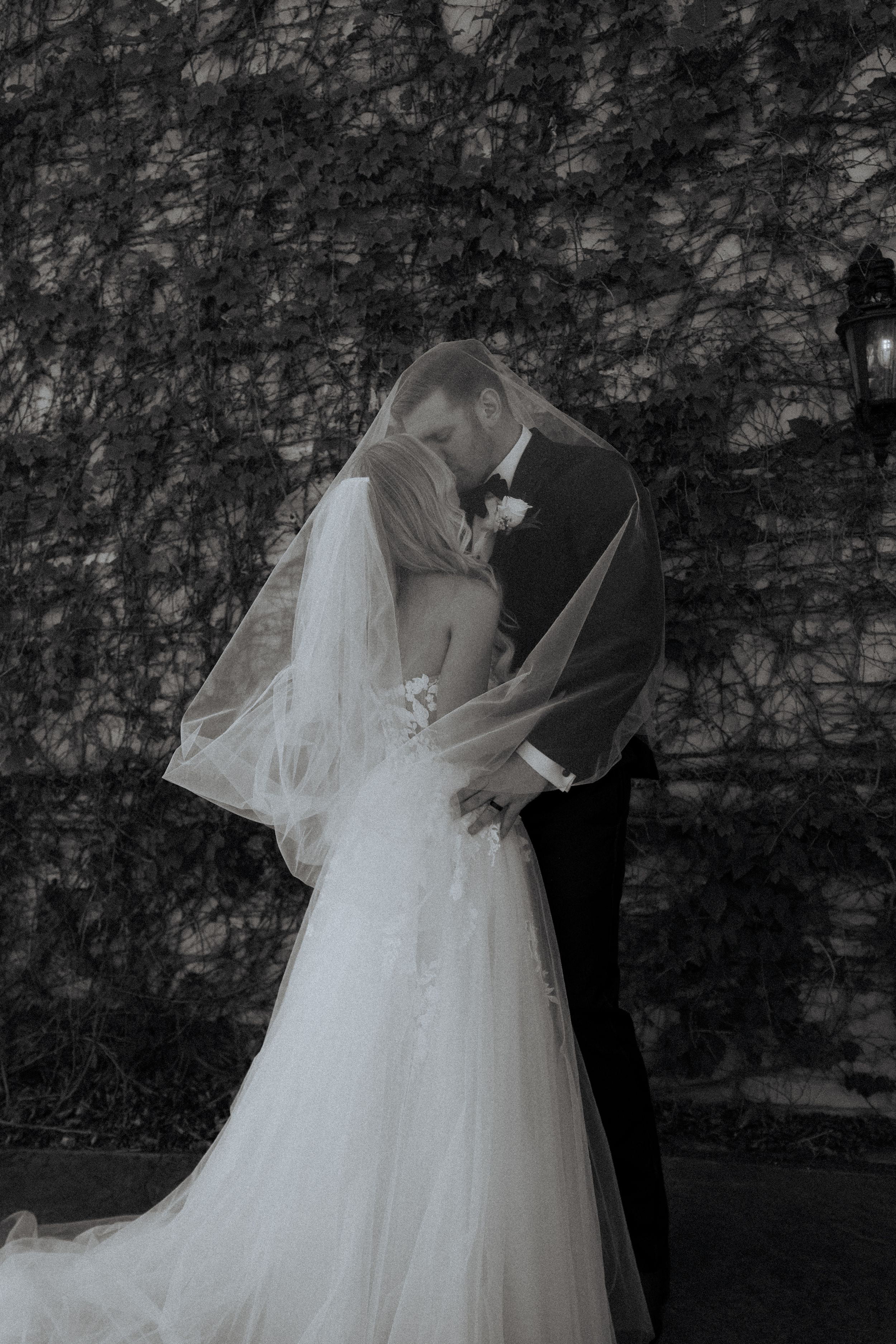 Black and white photo of a bride and groom sharing a kiss under her veil against an ivy-covered wall.