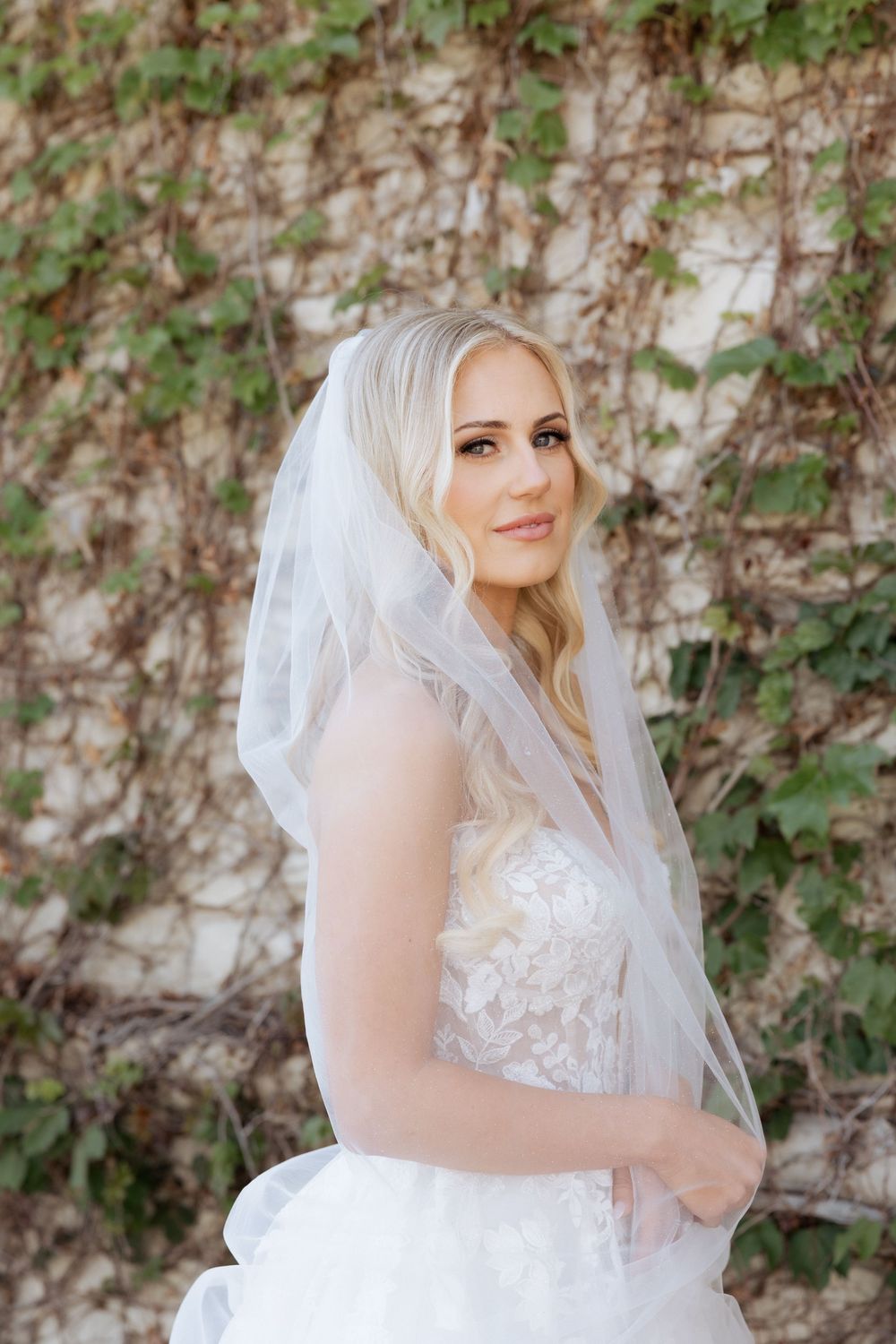 Blonde bride in white lace gown and veil poses gracefully against an ivy-covered stone wall.