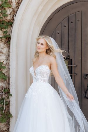 Blonde bride in strapless lace ballgown with flowing veil stands before arched stone doorway with dark wood door.