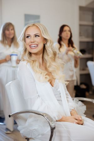 Smiling blonde bride in white robe sits in salon chair during wedding preparations with bridesmaids in background.