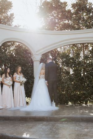 Bride and groom exchange vows under elegant white arch with bridesmaids in blush gowns at outdoor garden wedding ceremony.