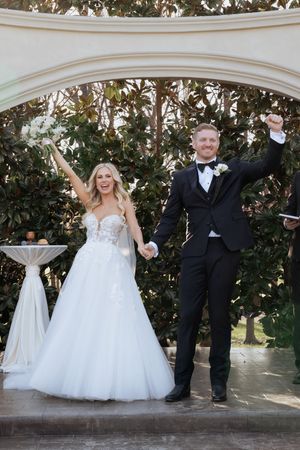 Joyful newlyweds celebrate with raised arms under white arch after outdoor garden wedding ceremony, bride in white gown.