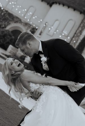 Black and white photo of groom dipping laughing blonde bride in wedding gown outdoors at venue.
