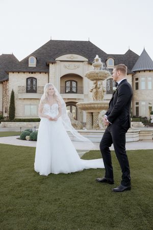 Bride and groom share a first look moment on lush green lawn before a grand stone mansion with elegant fountain.
