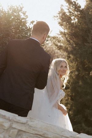Bride in lace wedding dress and veil smiles over shoulder at groom in black suit, surrounded by sunlit greenery.