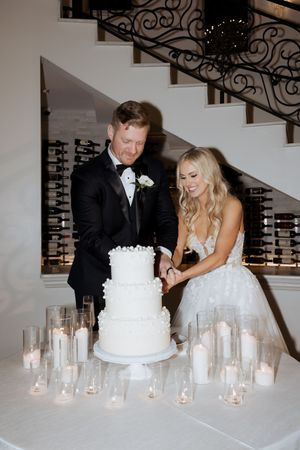Bride and groom cutting a three-tier white wedding cake surrounded by candles near an elegant staircase.