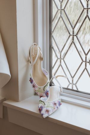 White ballet flats with floral embellishments sit elegantly on a windowsill with diamond-patterned glass panes.