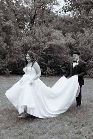A bride in a flowing white dress twirls and dances in a grassy field while a groom in black looks on.