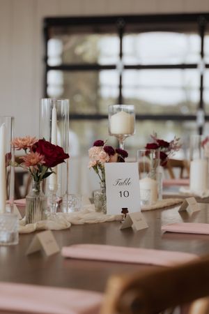 Elegant wedding reception table setting with burgundy flowers, candles and table number 10 card in soft natural lighting.