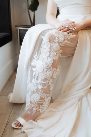 Close-up detail of white lace floral applique on sheer wedding dress sleeve with folded fabric on wooden floor.