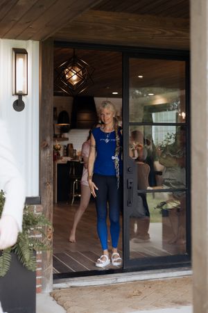 A person wearing a blue jumpsuit stands in a glass doorway with an exterior wall sconce lighting the entrance.