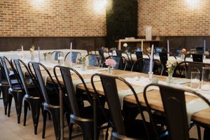 Modern restaurant interior with long wooden tables, black metal chairs, and warm lighting illuminating exposed brick walls.