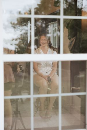 Through window panes, a person in white attire sits smiling indoors with a soft, dreamy look through the glass.