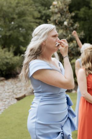 Woman in light blue dress standing outside during an event wipes her mouth with a napkin while another guest wears coral.