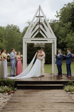 Wedding party poses playfully in sequence across white wooden chapel-style arches on rustic outdoor deck.