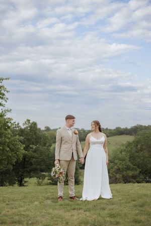 A couple stands together in a scenic meadow on their wedding day wearing a beige suit and flowing white dress.