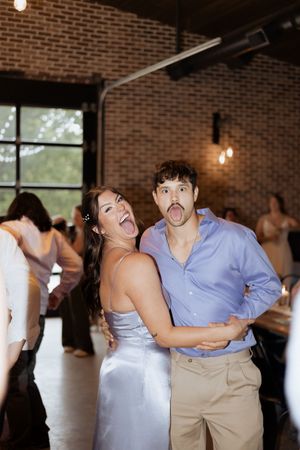 A couple makes silly faces while dancing together at a reception in a rustic brick-walled venue.