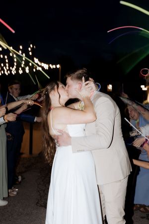 Newlyweds share a romantic kiss under twinkling string lights while wedding guests celebrate with sparklers at night.