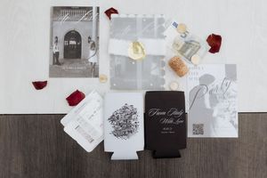 Wedding invitation suite with drink koozies, rose petals, and black and white design elements laid out on a wooden surface.