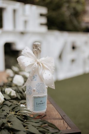 Glass bottles adorned with delicate white bows and lace details sit on a rustic wooden surface in an outdoor garden setting.