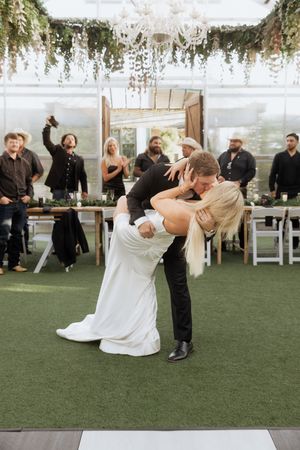 Wedding party dances in a greenhouse venue with hanging floral arrangements and white chairs on artificial turf.