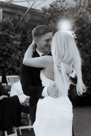 A black and white photograph captures a romantic moment between newlyweds dancing at their outdoor wedding reception.