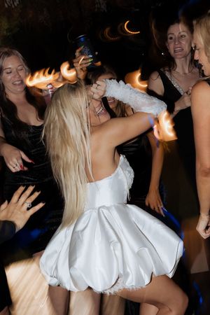Guests dance and celebrate on a dark dance floor as a woman in a short white bubble dress spins and dances.