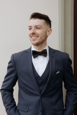 A groom wearing a classic black tuxedo with bow tie smiles in multiple candid poses against a light wall.