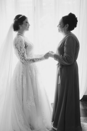 Silhouettes of two women stand near white curtains with one in a wedding dress and another in a dark gown.
