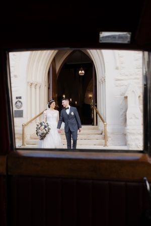 A wedding couple emerges through an arched stone church doorway with ornate architectural details.